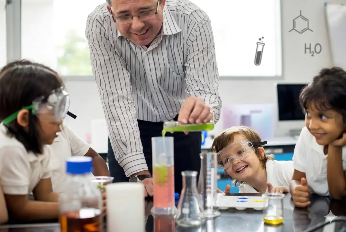 A teacher in a striped shirt and glasses pours a green liquid into a graduated cylinder of pink solution as a diverse group of excited young students in safety goggles watches attentively around a lab table with beakers and flasks.