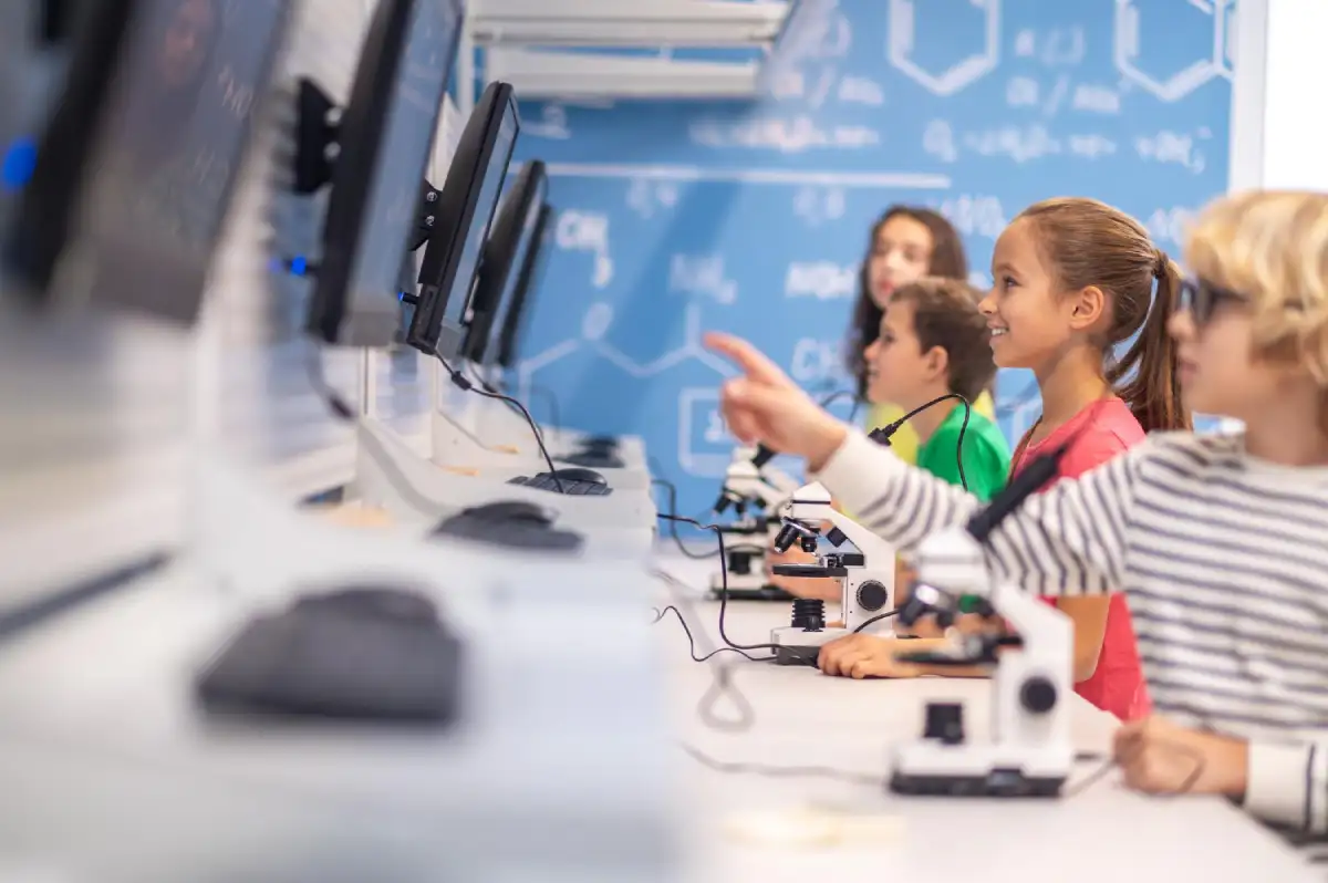 A group of diverse elementary school children smiling and engaged in a science lab. They are seated at a long white desk equipped with microscopes and computer monitors, while the background features a blue wall covered in white chemical formulas and molecular structures.