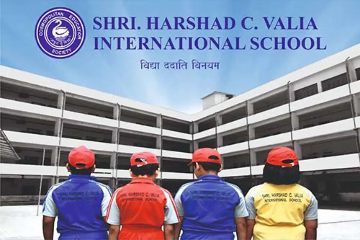 Students standing in front of Shri Harshad C. Valia International School campus wearing colorful school uniforms and caps.